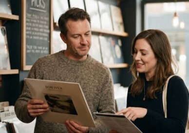 Un hombre de mediana edad con un suéter de punto marrón mira pensativamente un disco de vinilo de Bon Iver que sostiene en sus manos. Está de pie junto a una vendedora con un suéter negro que sonríe y señala información en un folleto. Están dentro de una acogedora tienda de discos con estantes de madera llenos de vinilos, paredes de color carbón oscuro, lámparas de cobre de estilo industrial y un letrero que dice "Staff Picks" (Selecciones del personal) en el fondo